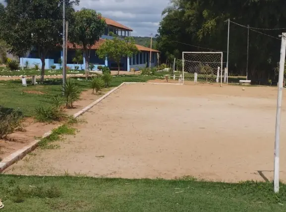Campo de terra batida com uma trave ao fundo, gramado em volta e um sobrado azul à esquerda